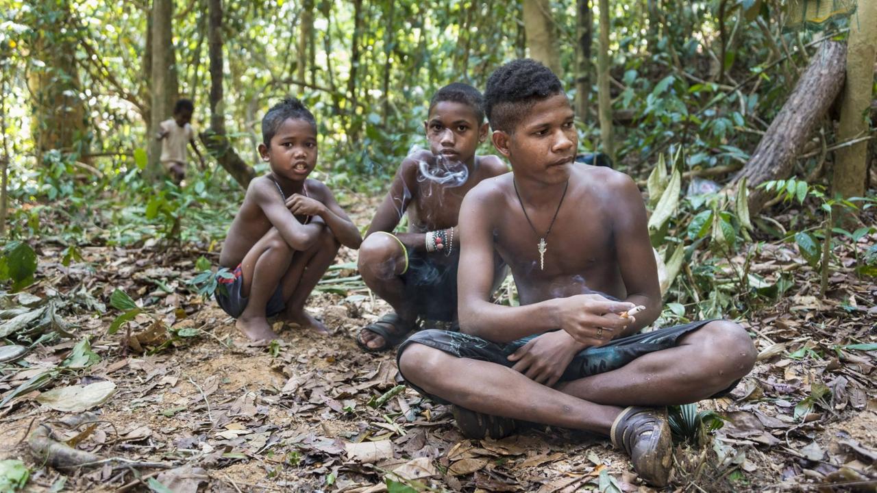 Drei Jungen der Orang Asli sitzen auf dem Boden im Dschungel und rauchen Drei Jungen der Orang Asli sitzen auf dem Boden im Dschungel und rauchen
