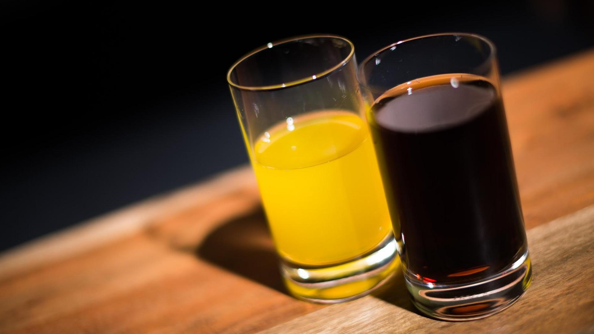 Ein Glas mit Cola steht neben einem Glas mit Orangenlimonade. Ein Glas mit Cola steht neben einem Glas mit Orangenlimonade.
