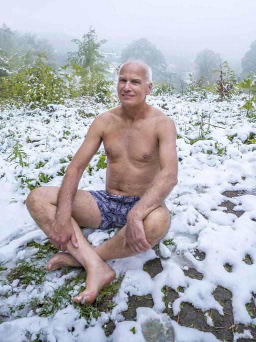 Ein älterer Mann sitzt nur in Unterhose auf einer schneebedeckten Wiese.