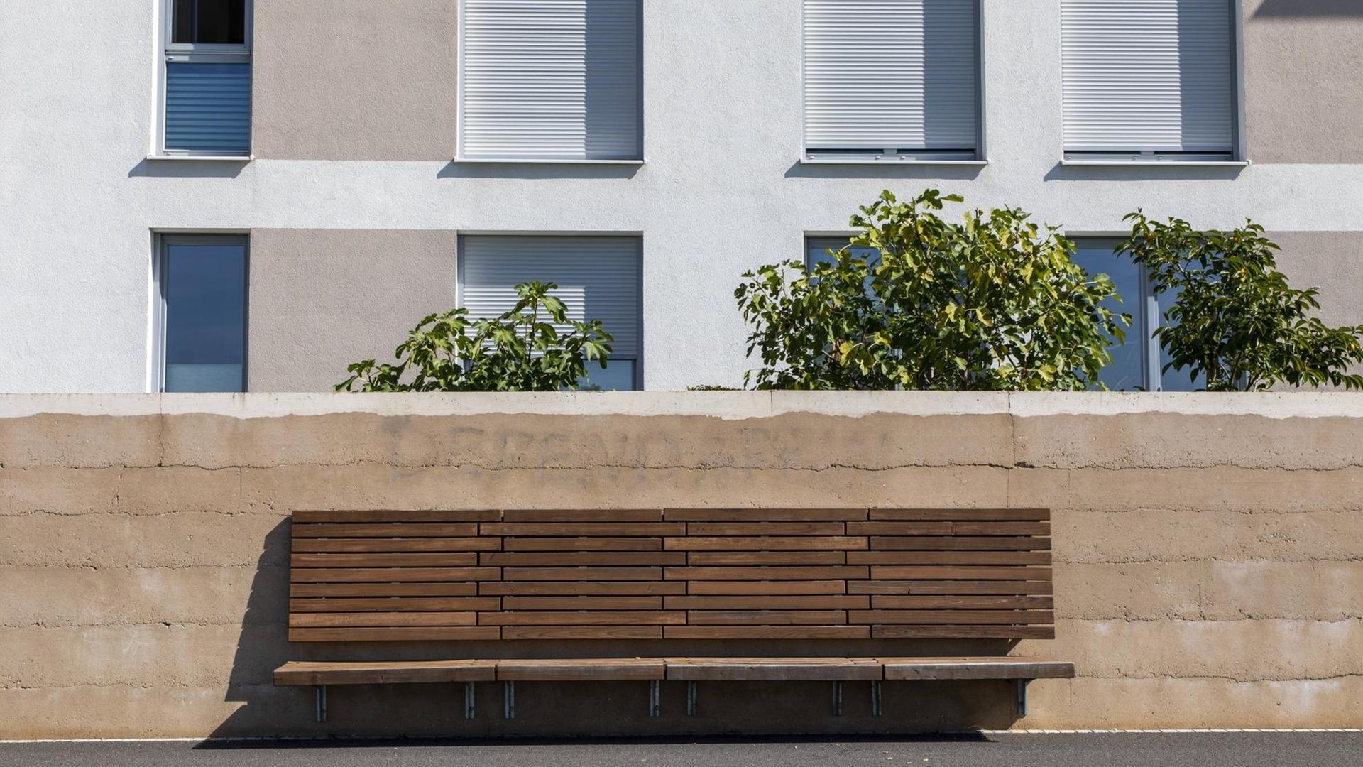 Eine leere Bank steht vor einer Gartenmauer, dahinter ein Reihenhaus mit von Jalousien verschlossenen Fenstern. Eine leere Bank steht vor einer Gartenmauer, dahinter ein Reihenhaus mit von Jalousien verschlossenen Fenstern.