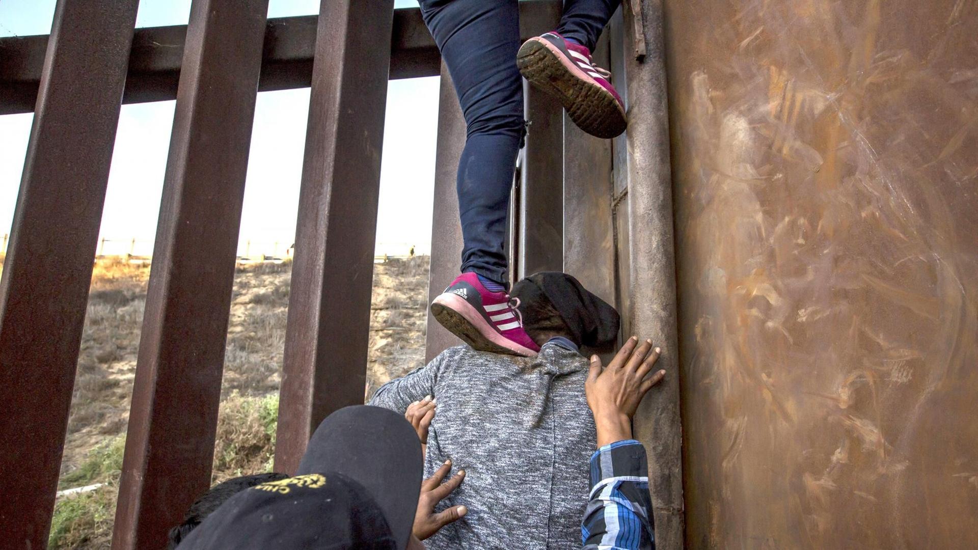 Migranten aus Mexiko klettern über den Grenzzaun in die USA. Füsse stehen auf einer Schulter und Hände werden sichernd in die Luft gereckt. Migranten aus Mexiko klettern über den Grenzzaun in die USA. Füsse stehen auf einer Schulter und Hände werden sichernd in die Luft gereckt.