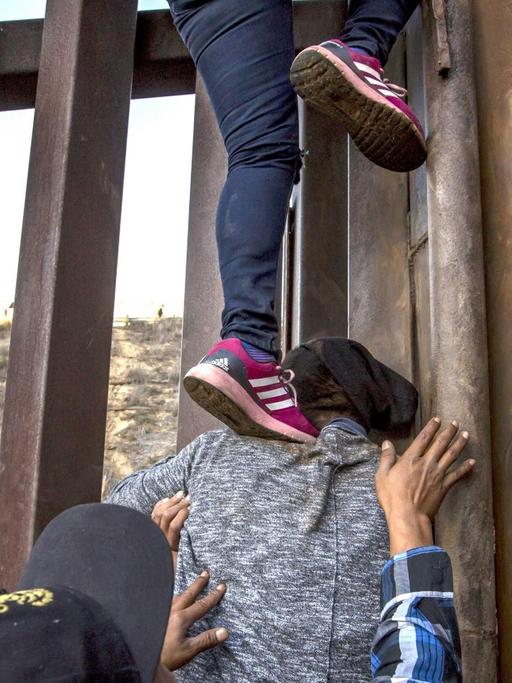Migranten aus Mexiko klettern über den Grenzzaun in die USA. Füsse stehen auf einer Schulter und Hände werden sichernd in die Luft gereckt.