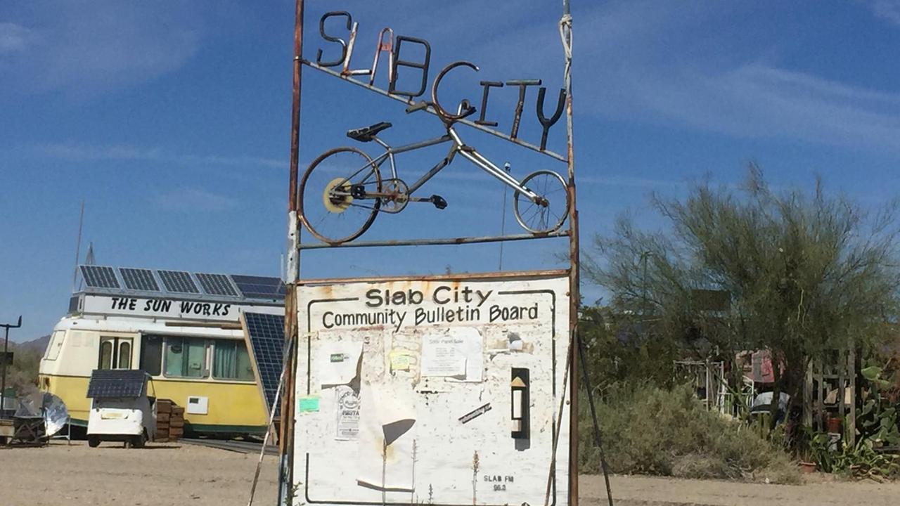Ein rostiges Schild mit dem Schriftzug "Slab City" steht im Wüstensand. Dahinter ein gelber Bus mit alten Solar-Panelen.