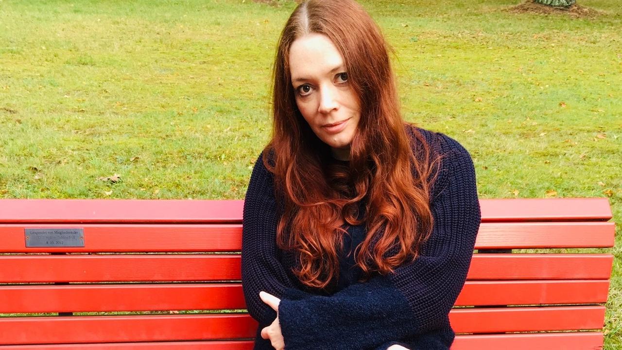 Zoë Beck sitzt auf einer roten Parkbank. Hinter ihr ist Rasen. Zoë Beck sitzt auf einer roten Parkbank. Hinter ihr ist Rasen.