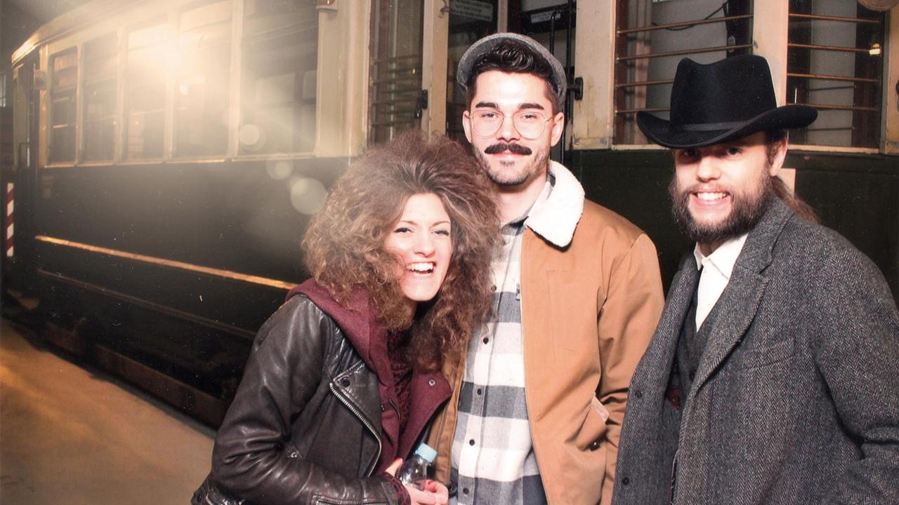 Eine Frau mit blonden lockigen Haaren und zwei Männer stehen vor einem historischen Zug. Der Mann rechts trägt einen schwarzen Hut.