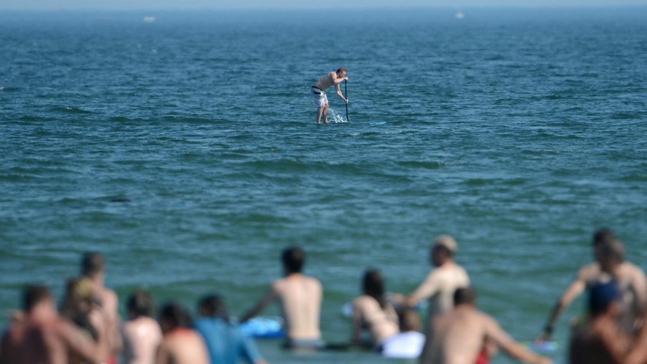 Ein Mann steht auf einem Paddelbrett etwa 100 m vor der Küste, während Menschen am Nauset Beach auf Cape Cod in Orleans, Massachusetts, USA baden. 