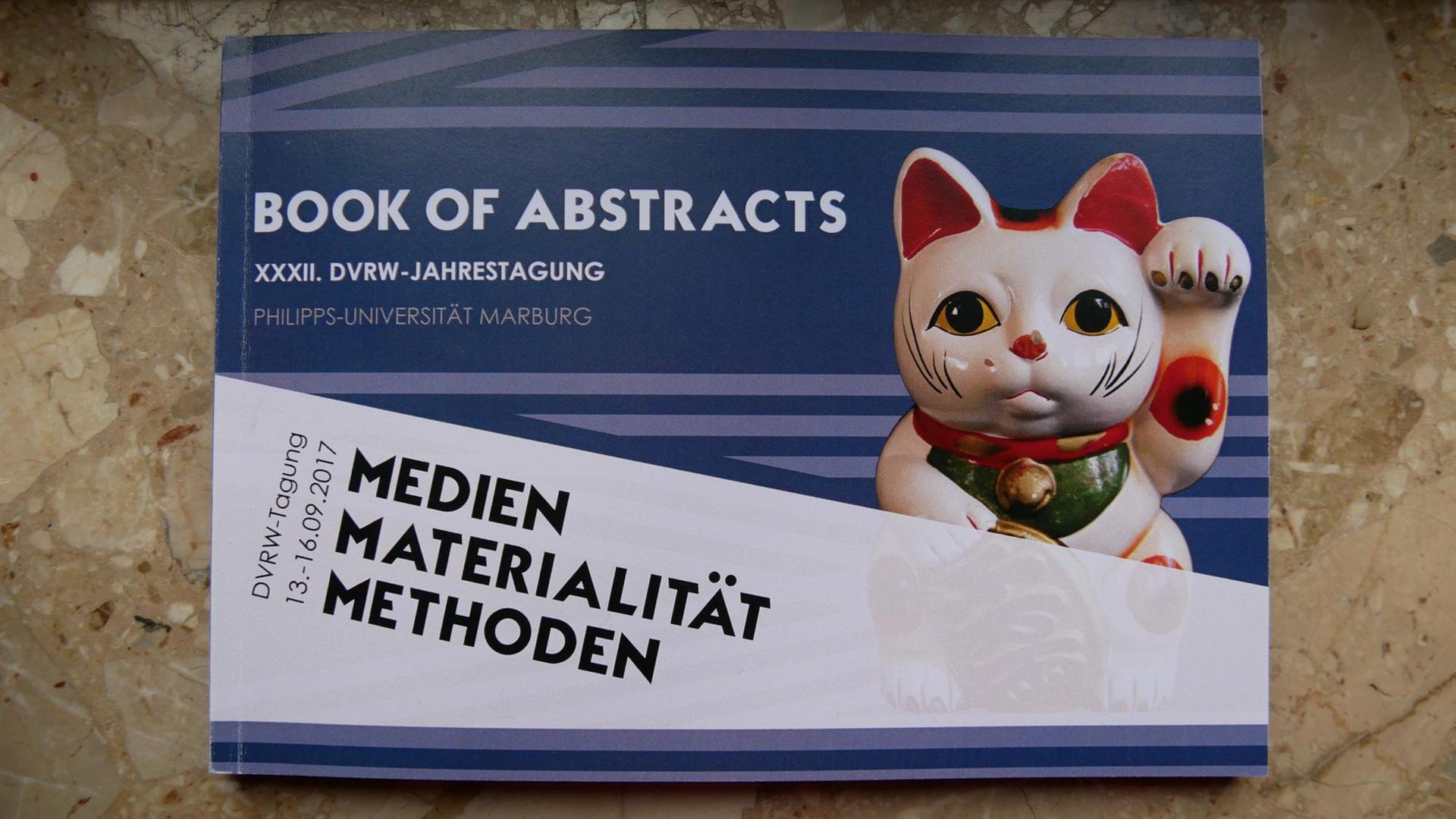 Das Programmheft der diesjährigen Tagung der Religionswissenschaften an der Universität Marburg - darauf abgebildet ist eine Winkekatze Das Programmheft der diesjährigen Tagung der Religionswissenschaften an der Universität Marburg - darauf abgebildet ist eine Winkekatze
