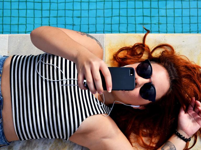 Eine junge Frau liegt am Rand eines Pools und kommunziert über ihr Handy.