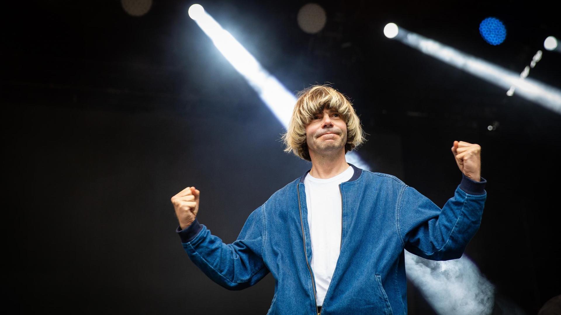 Der Musiker Tim Burgess steht in einer blauglänzenden Jacke auf einer Bühne, im Hintergrund Scheinwerferlicht. Der Musiker Tim Burgess steht in einer blauglänzenden Jacke auf einer Bühne, im Hintergrund Scheinwerferlicht.
