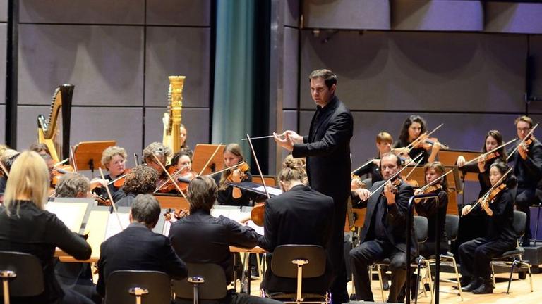 Der Dirigent steht vor dem Orchester und blickt Richtung der ersten Geigen. Der Dirigent steht vor dem Orchester und blickt Richtung der ersten Geigen.