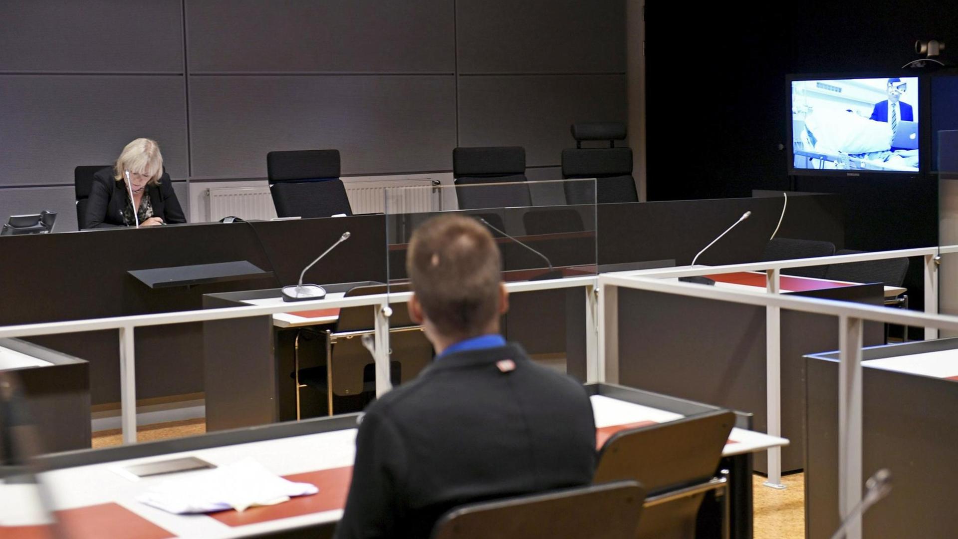 Das Foto zeigt einen abgedunkelten Gerichtssaal mit einem Bildschirm, auf dem ein Tatverdächtiger per Videoübertragung an der Gerichtssitzung teilnimmt. Das Foto zeigt einen abgedunkelten Gerichtssaal mit einem Bildschirm, auf dem ein Tatverdächtiger per Videoübertragung an der Gerichtssitzung teilnimmt.