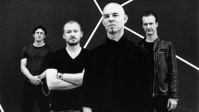 Vier in schwarz gekleidete Männer stehen versetzt aufgestellt vor einem schwarzem Hintergrund mit strahlenartigem Muster.