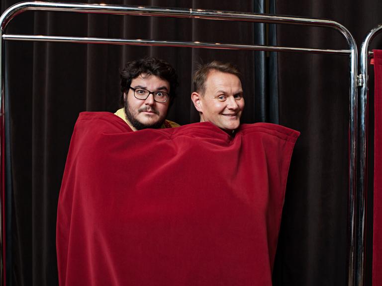 Das Bild zeigt Axel Ranisch und Devid Striesow, die sich zusammen in einen roten Vorhang eingewickelt haben.