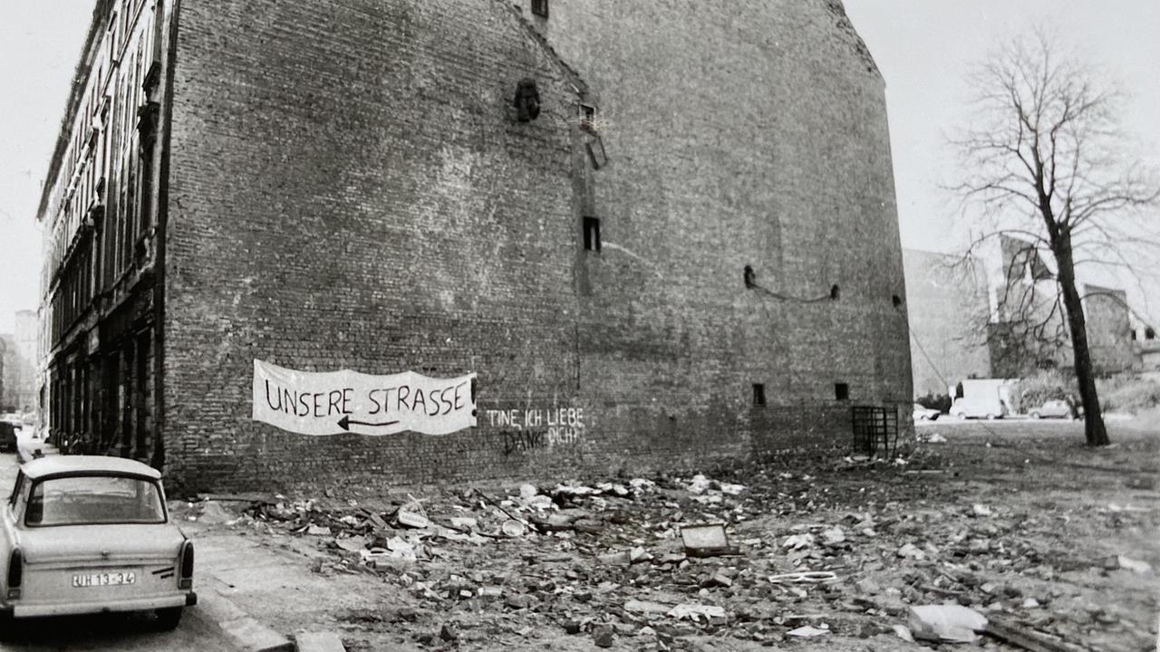 Schwarz-Weiß-Bild eines verfallenen Hause neben einer Brache mit Schutt. An der Fassade steht „Unsere Straße“. Schwarz-Weiß-Bild eines verfallenen Hause neben einer Brache mit Schutt. An der Fassade steht „Unsere Straße“.