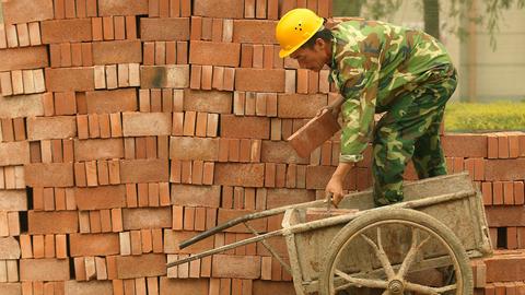 Chinesischer Bauarbeiter stapelt Backsteine in Peking. Er steht mit einem Bein in einer Schubkarre