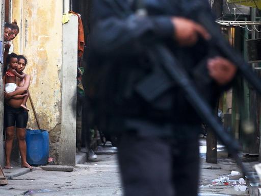 In der Favela Maré in der brasilianische Rio de Janeiro schauen drei schwarze Frauen und ein Kind aus einem Hauseinagang auf einen mit einem Gewehr bewaffenten Polizisten.