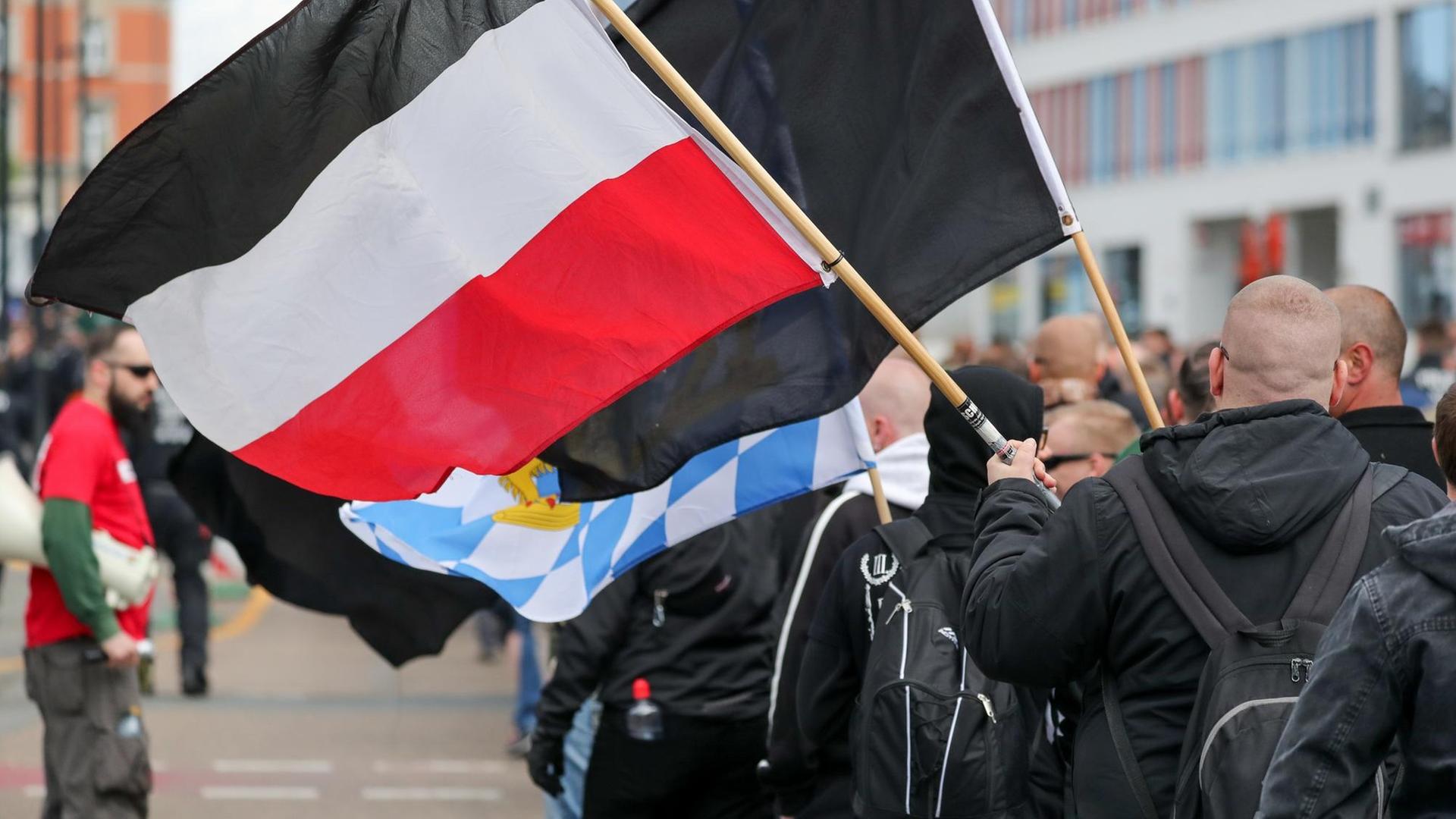 Glatzköpfige, in schwarz gekleidete Männer sind von hinten zu sehen, wie sie mit Flaggen durch die Straße ziehen. Glatzköpfige, in schwarz gekleidete Männer sind von hinten zu sehen, wie sie mit Flaggen durch die Straße ziehen.