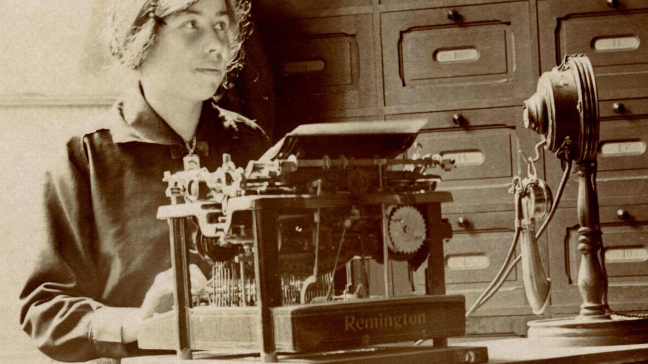 Schwarzweißfoto einer Sekretärin an ihrem Schreibtisch vor einer Remington-Schreibmaschine und ihrem Telefon - die Geräte wirkten ihrem Alter entsprechend sehr altertümlich.