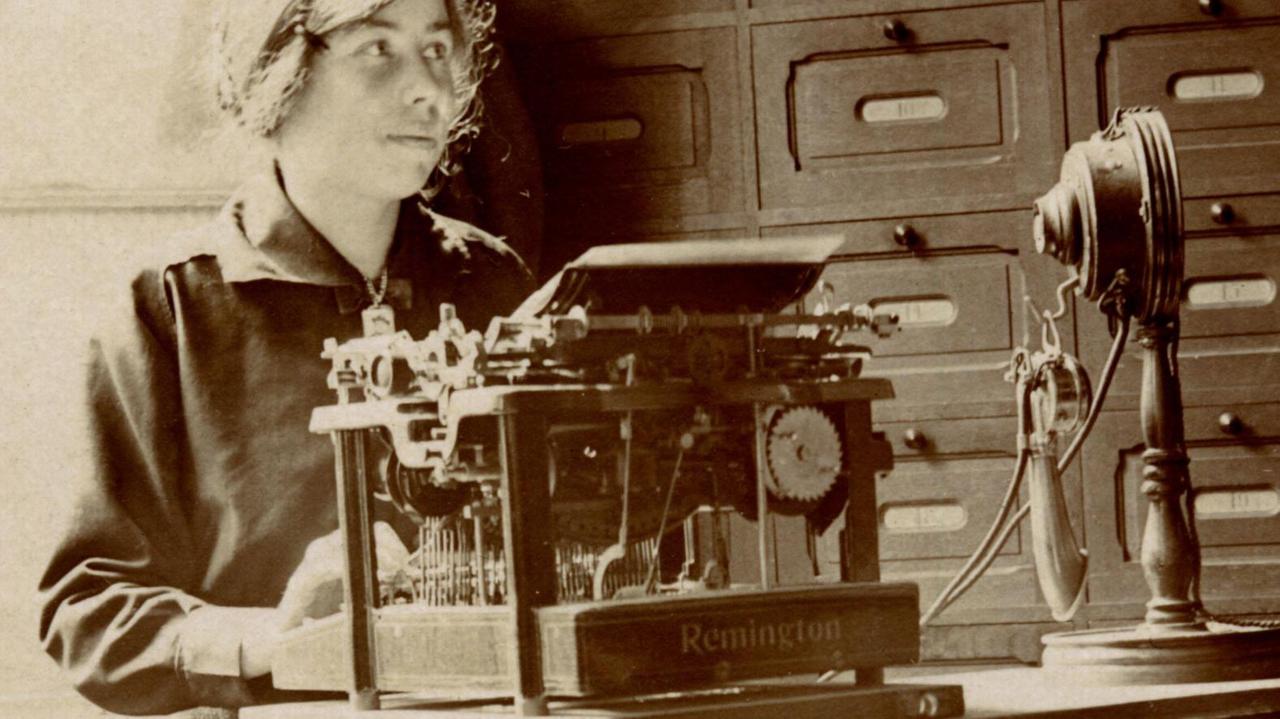 Schwarzweißfoto einer Sekretärin an ihrem Schreibtisch vor einer Remington-Schreibmaschine und ihrem Telefon - die Geräte wirkten ihrem Alter entsprechend sehr altertümlich. Schwarzweißfoto einer Sekretärin an ihrem Schreibtisch vor einer Remington-Schreibmaschine und ihrem Telefon - die Geräte wirkten ihrem Alter entsprechend sehr altertümlich.