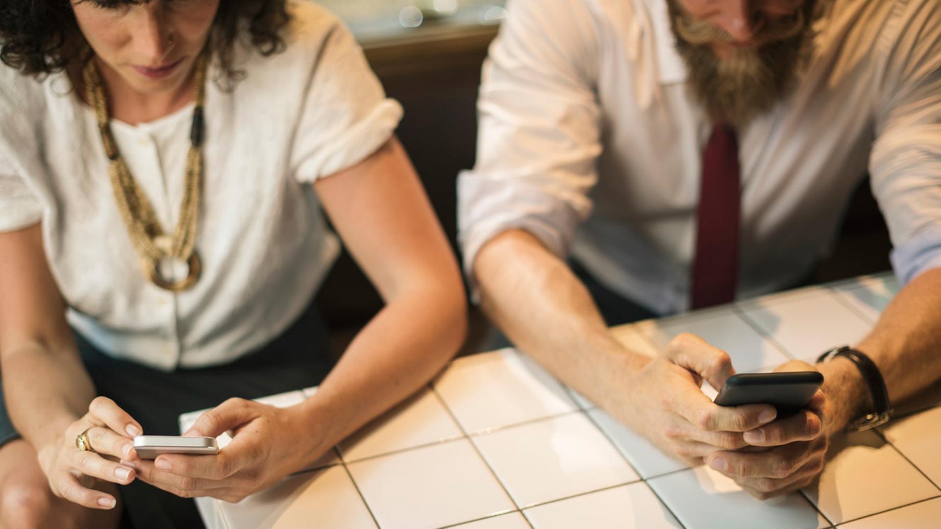 Eine Frau und ein Mann sitzen nebeneinander und kommunizieren jeder für sich über ihr Smartphone. Eine Frau und ein Mann sitzen nebeneinander und kommunizieren jeder für sich über ihr Smartphone.