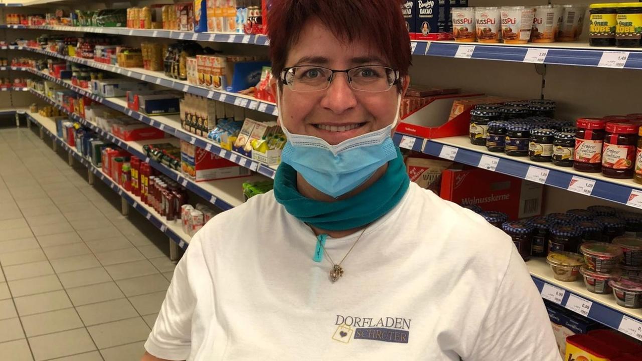 Silke Schröter steht mit Mundschutz und weißem T-Shirt vor den Regalen ihres Dorfladens. Silke Schröter steht mit Mundschutz und weißem T-Shirt vor den Regalen ihres Dorfladens.