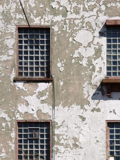 Fassade mit sich lösendem Putz und teilweise kaputten Glasfliesenfenstern.