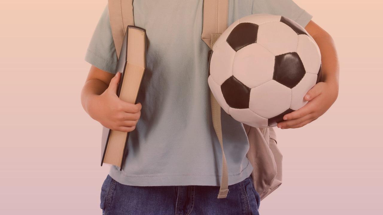 Eine Studioaufnahme zeigt einen kleinen Jungen im Anschnitt mit einem Buch unter dem rechten und einem Fußball unter dem linken Arm. Eine Studioaufnahme zeigt einen kleinen Jungen im Anschnitt mit einem Buch unter dem rechten und einem Fußball unter dem linken Arm.