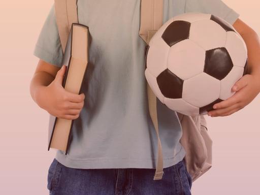 Eine Studioaufnahme zeigt einen kleinen Jungen im Anschnitt mit einem Buch unter dem rechten und einem Fußball unter dem linken Arm.