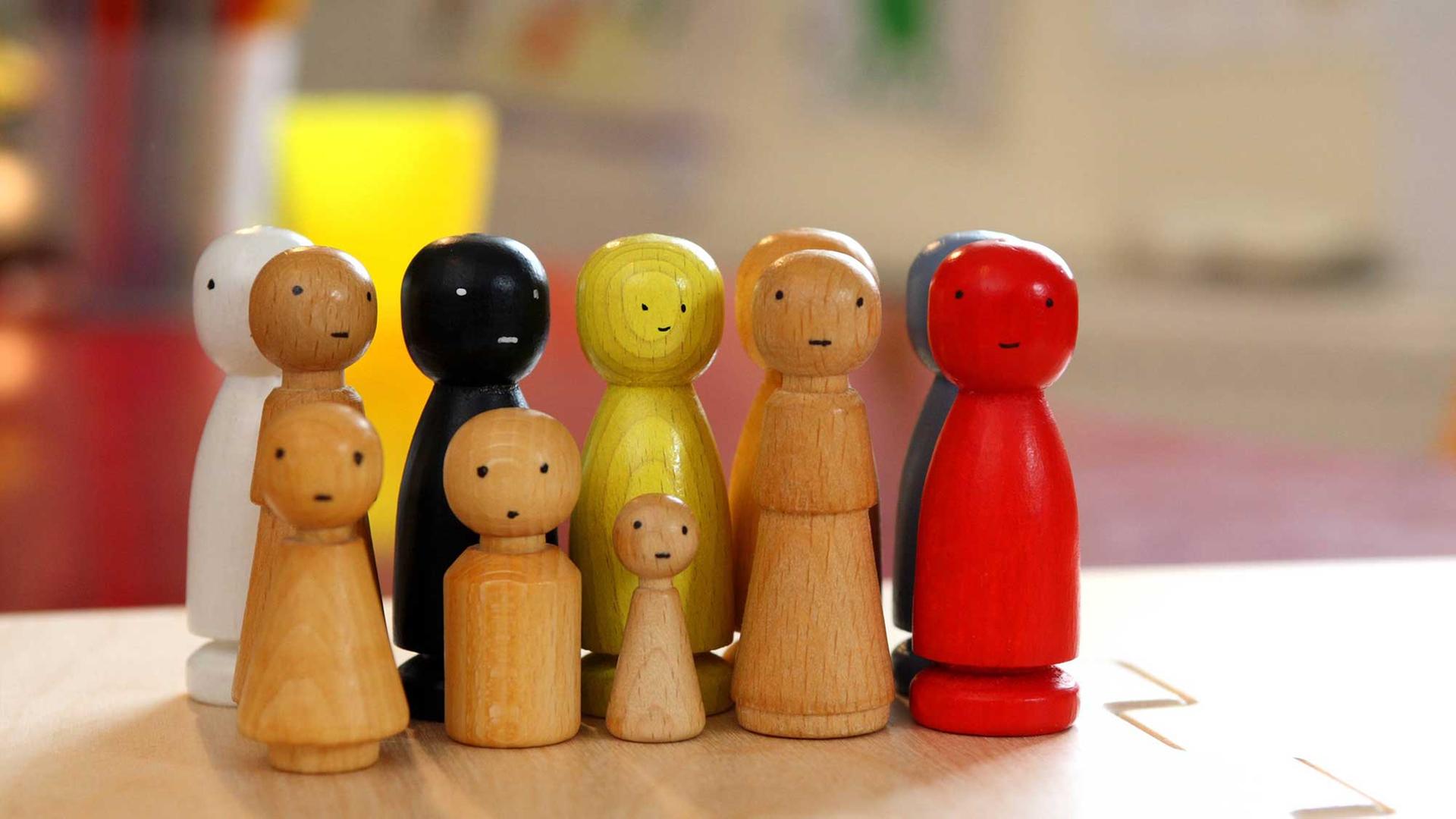 Mehrere wie Menschen aussehende Holzfiguren stehen auf einem Tisch, einige davon sind farbig angemalt, die Größen der Figuren unterscheiden sich