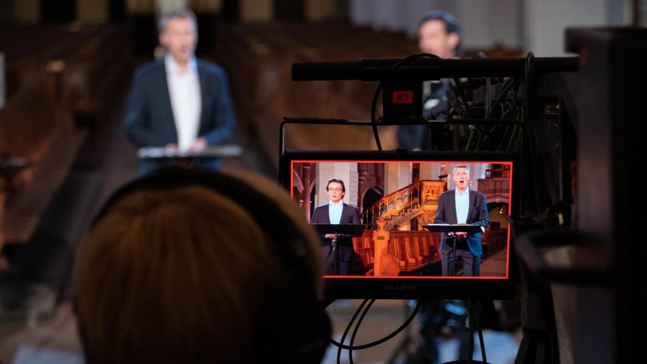 Ein Mann schaut über einen Monitor auf die Szene, die er gerade filmt: einen Sänger, der hinter einem Notenständer steht. Ein Mann schaut über einen Monitor auf die Szene, die er gerade filmt: einen Sänger, der hinter einem Notenständer steht.