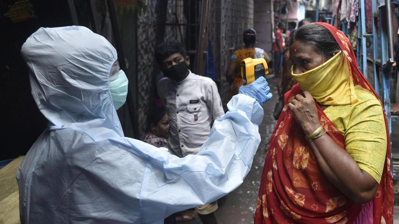 Ein Mann im weißen Schutzanzug misst in einer engen Gasse bei einer Frau im rot-gelben Sari die Temperatur.