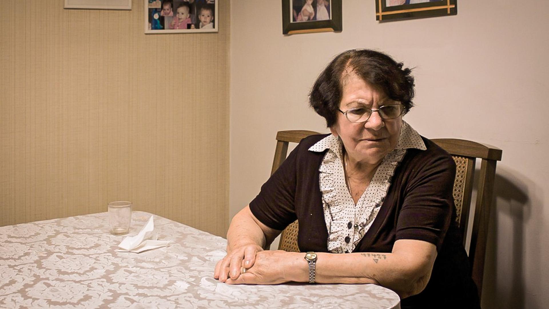 Die Shoah-Überlebende Frieda Kliger sitzt an ihrem Küchentisch und blickt nachdenklich. Auf Ihrem linken Arm ist eine tätowierte Nummer erkennbar.
