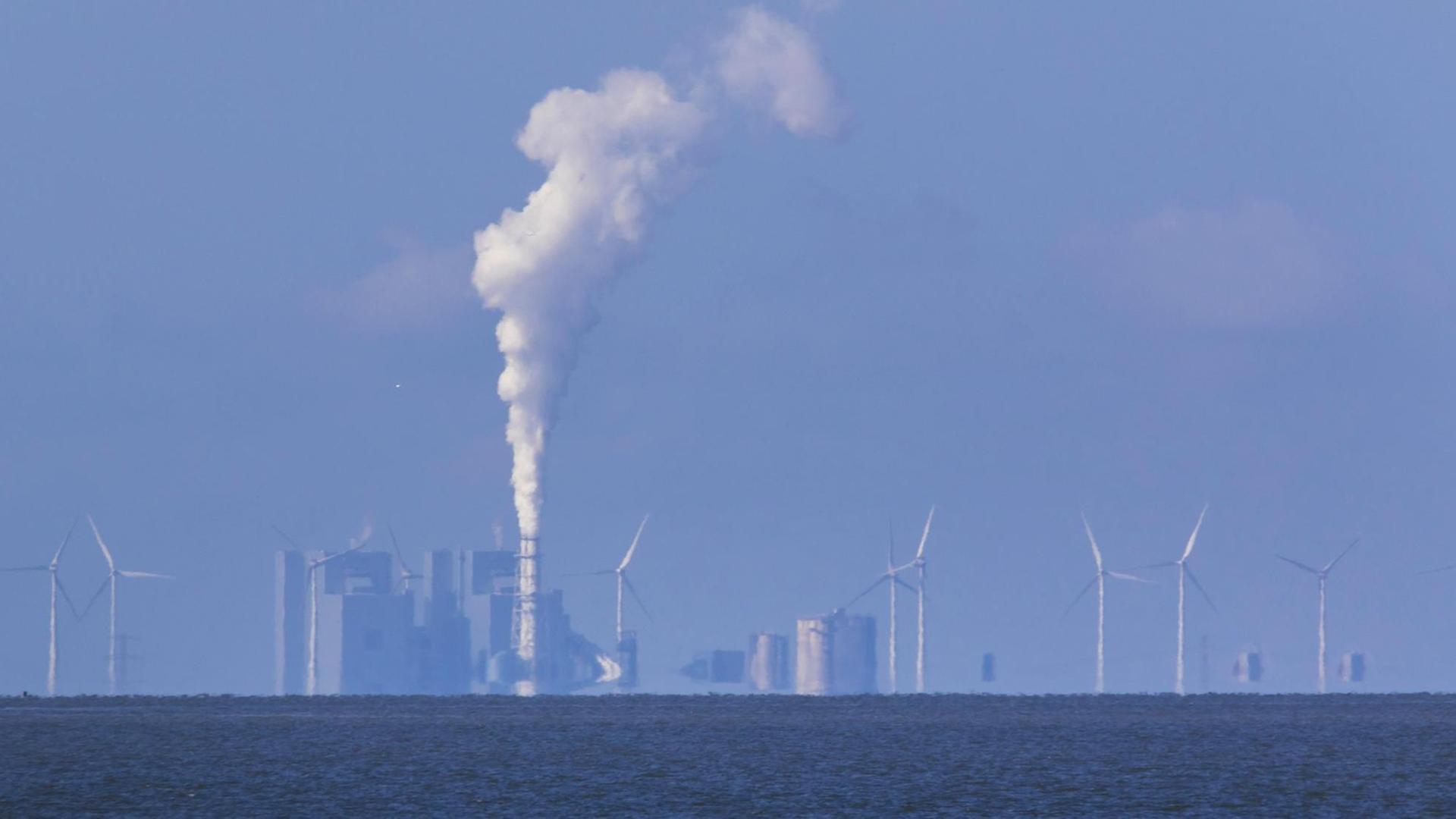 Blick aufs Meer, an dessen Horizont sich ein industrieller Komplex aus Wind- und Kohlekraft zeigt. Weißer Rauch steigt auf.