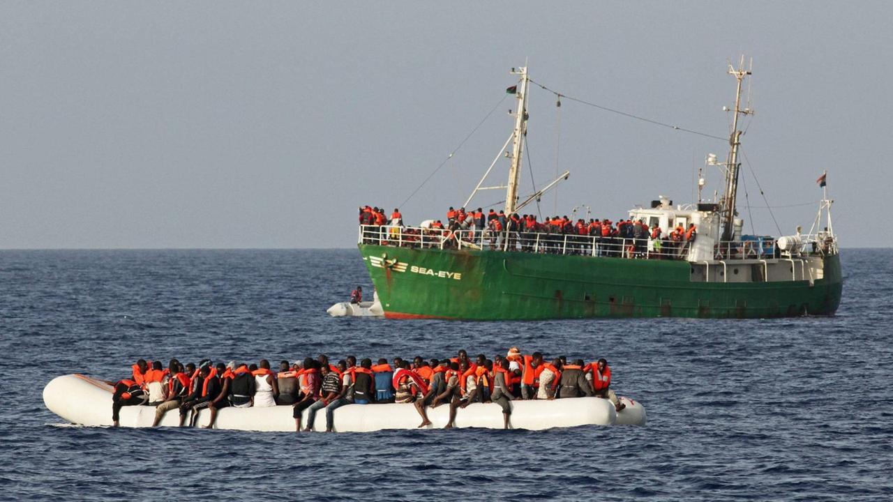 Im Vordergrund ein überfülltes Schlauchboot mit Menschen. Im Hintergrund die "Sea Eye" ein Seenotrettungsschiff. Im Vordergrund ein überfülltes Schlauchboot mit Menschen. Im Hintergrund die "Sea Eye" ein Seenotrettungsschiff.