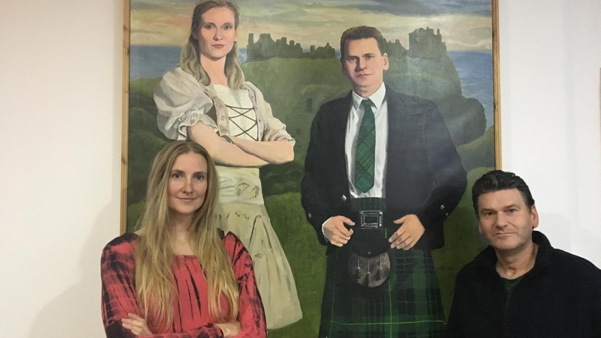 Das Künstlerpaar Bibo und Brain Keeley vor einem Gemälde mit beiden in schottischer Tracht