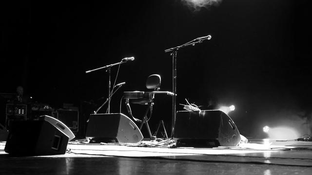 Schwarz-weißes Bild einer menschenleeren Bühne mit Monitorboxen, Mikrofonen auf Stativen und Gitarren.