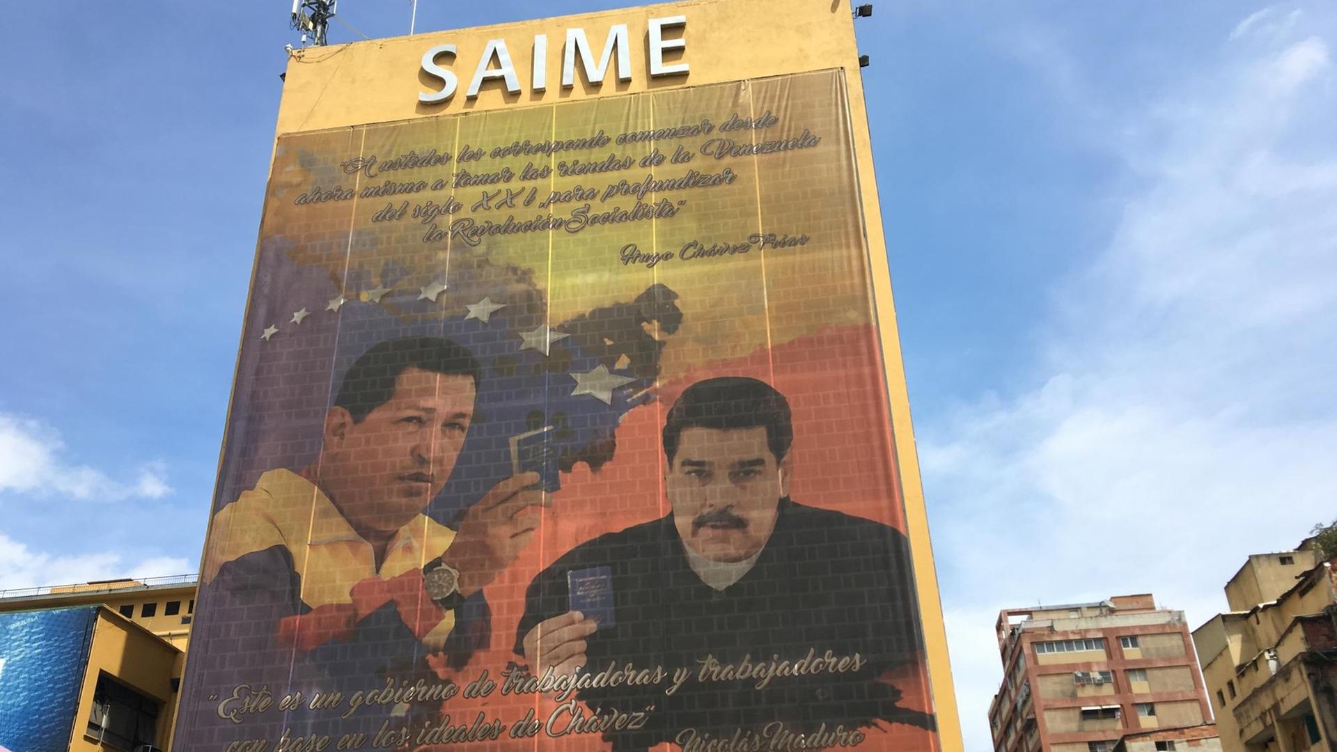 Auf einer Fassadenplane sind Hugo Chavez und Nicolas Maduro zu sehen, darüber steht das Wort SAIME.