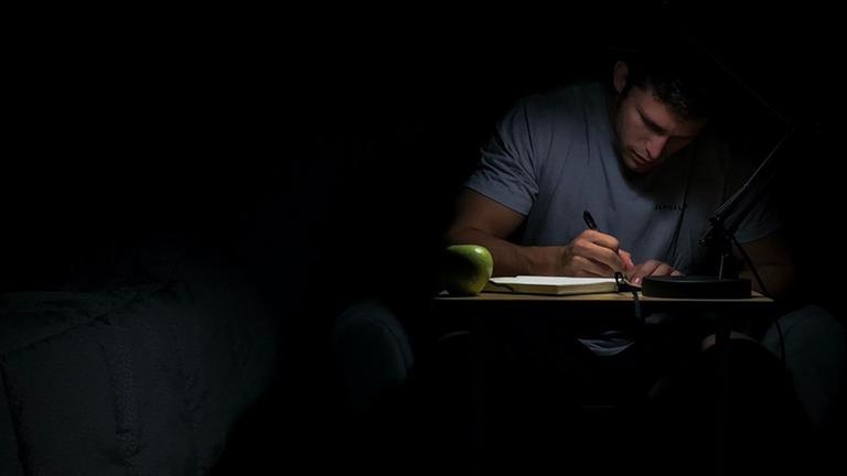 Ein Mann sitzt in einem dunklen Zimmer und schreibt im Schein einer zielgerichteten Lampe mit der Hand in einen Block, Ein Mann sitzt in einem dunklen Zimmer und schreibt im Schein einer zielgerichteten Lampe mit der Hand in einen Block,