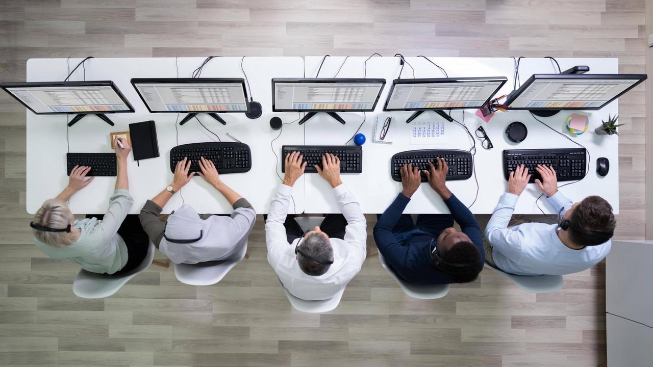 Draufsicht auf einen weißen Schreibtisch mit diversen Computerbildschirmen vor denen eng nebeneinander fünf Menschen arbeiten Draufsicht auf einen weißen Schreibtisch mit diversen Computerbildschirmen vor denen eng nebeneinander fünf Menschen arbeiten