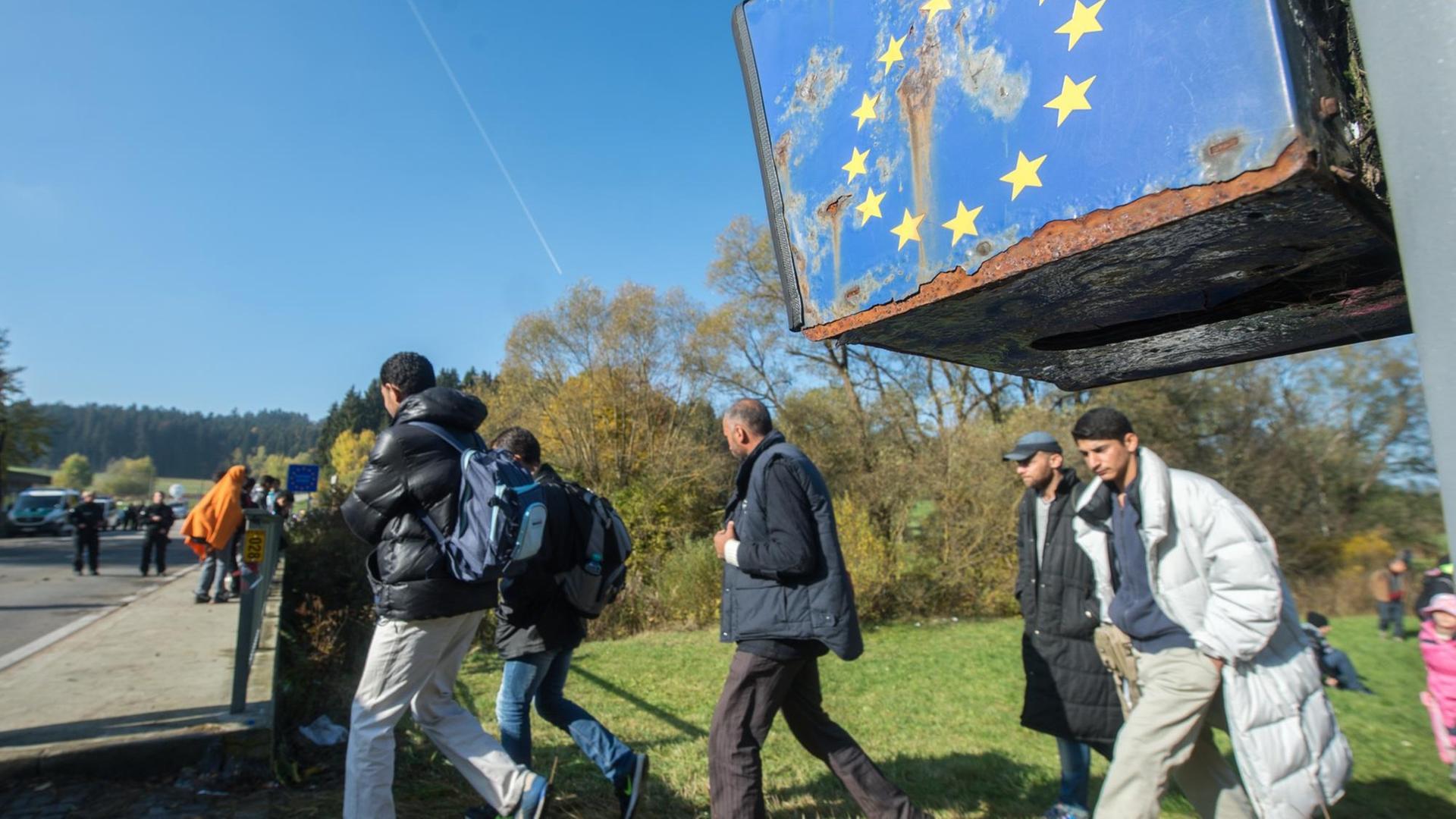 Flüchtlinge laufen über ein Feld auf eine Straße, unter einem ramponierten EU-Logo.