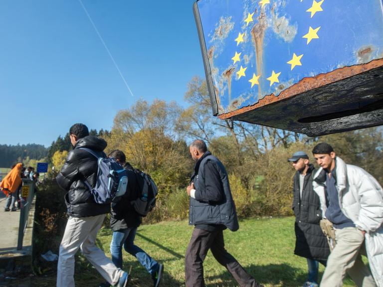 Flüchtlinge laufen über ein Feld auf eine Straße, unter einem ramponierten EU-Logo.