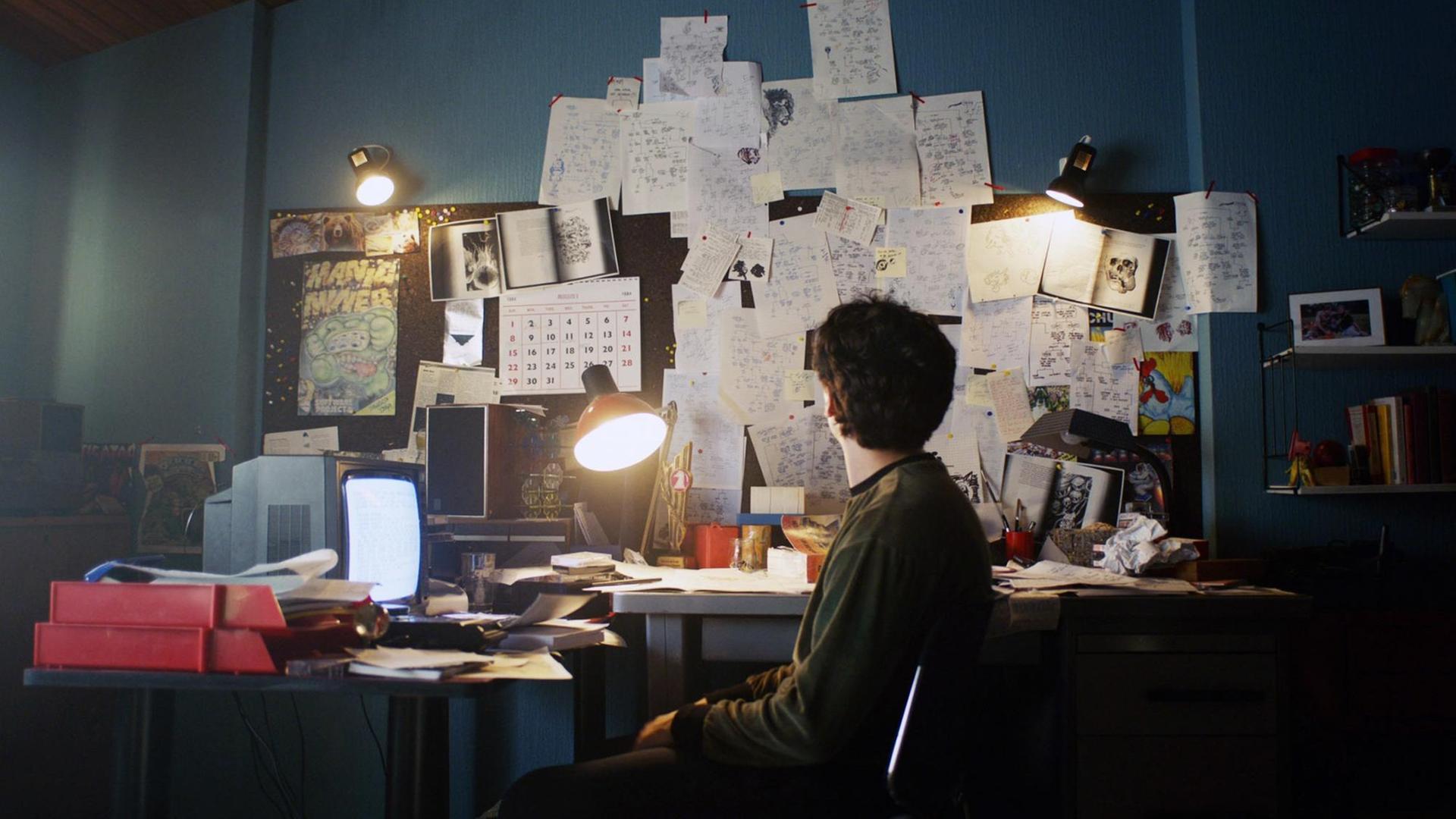 Filmstill aus der Netflix Serie "BLACK MIRROR: BANDERSNATCH. Der Schauspieler Fionn Whitehead in der Hauptrolle sitzt vor einem Bildschirm an einem chaotischem Schreibtisch. Filmstill aus der Netflix Serie "BLACK MIRROR: BANDERSNATCH. Der Schauspieler Fionn Whitehead in der Hauptrolle sitzt vor einem Bildschirm an einem chaotischem Schreibtisch.