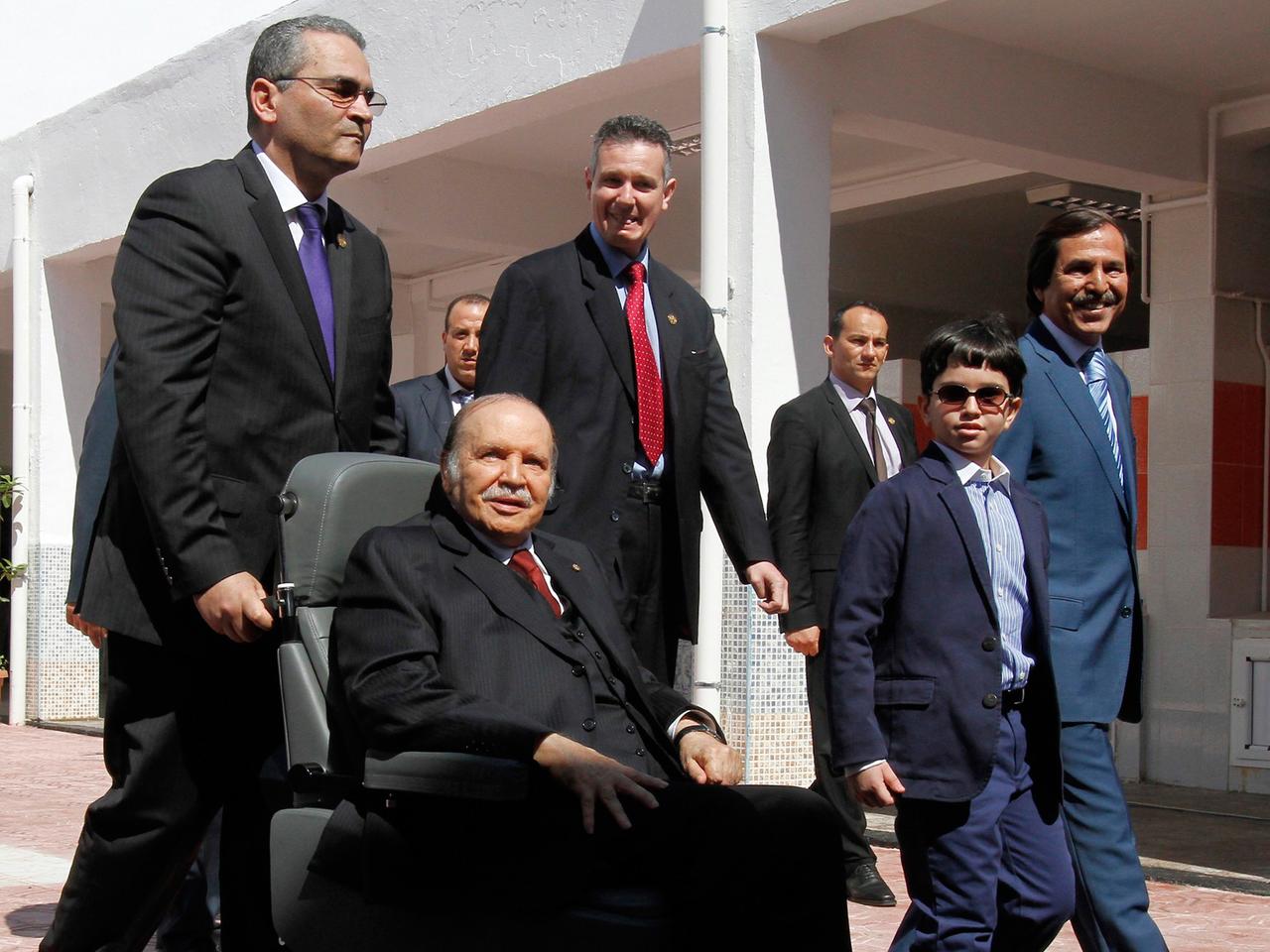 Abdelaziz Bouteflika wird im Rollstuhl von einem Mann geschoben, hinter ihm weitere Männer und ein Junge Abdelaziz Bouteflika wird im Rollstuhl von einem Mann geschoben, hinter ihm weitere Männer und ein Junge