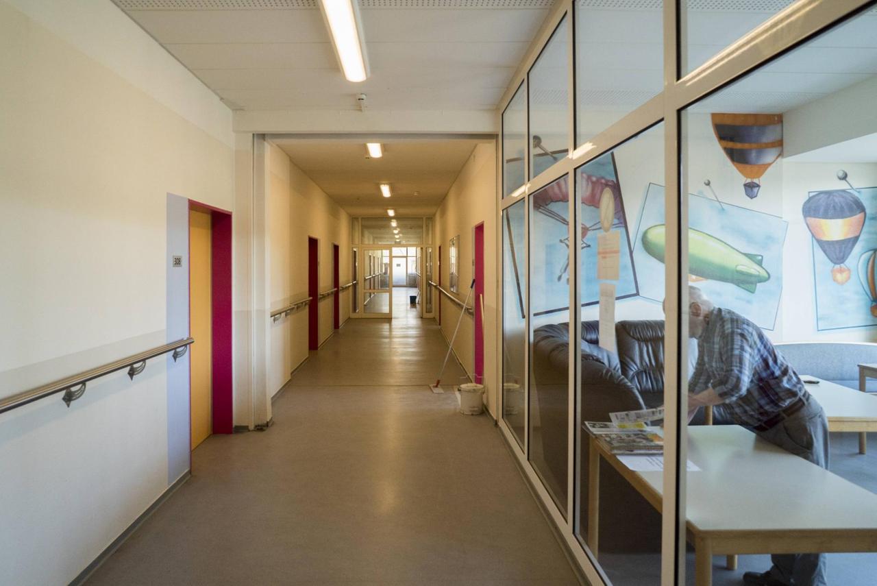 Ein langer Gang mit handläufen an der Wand. Rechts ist ein Gemeinschaftsraum zu sehen in dem ein älterer Herr an einem Tisch steht. Ein langer Gang mit handläufen an der Wand. Rechts ist ein Gemeinschaftsraum zu sehen in dem ein älterer Herr an einem Tisch steht.