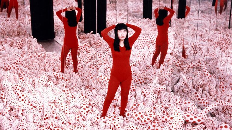 Eine Installation der japanischen Künstlerin Yayai Kusama. Eine Frau steht in einem Feld phallusförmiger, weiß-rot-gemusterter Gebilde, die den Boden bedecken. Das Feld ist von Spiegeln umgeben. Eine Installation der japanischen Künstlerin Yayai Kusama. Eine Frau steht in einem Feld phallusförmiger, weiß-rot-gemusterter Gebilde, die den Boden bedecken. Das Feld ist von Spiegeln umgeben.