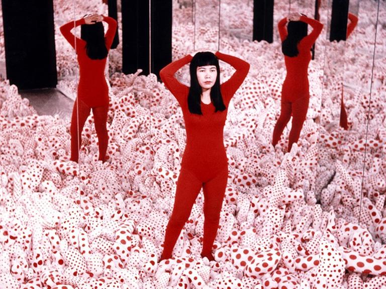 Eine Installation der japanischen Künstlerin Yayai Kusama. Eine Frau steht in einem Feld phallusförmiger, weiß-rot-gemusterter Gebilde, die den Boden bedecken. Das Feld ist von Spiegeln umgeben.