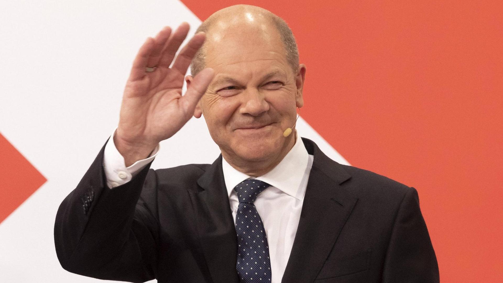 Der künftige Bundeskanzler Olaf Scholz steht in einem schwarzen Anzug vor einer rot-weißen Wand und winkt. Der künftige Bundeskanzler Olaf Scholz steht in einem schwarzen Anzug vor einer rot-weißen Wand und winkt.