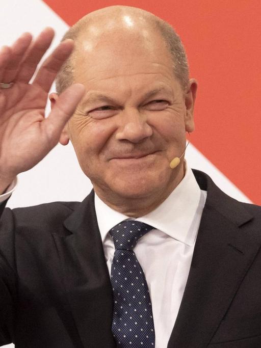 Der künftige Bundeskanzler Olaf Scholz steht in einem schwarzen Anzug vor einer rot-weißen Wand und winkt.