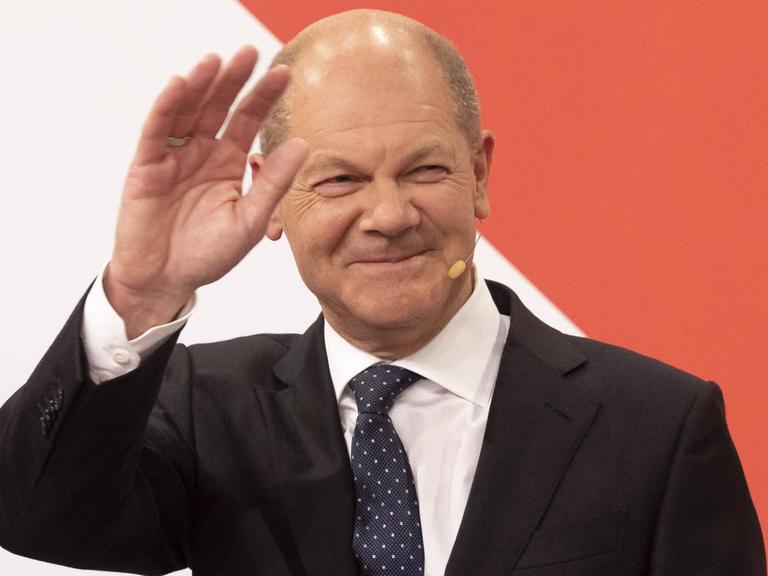 Der künftige Bundeskanzler Olaf Scholz steht in einem schwarzen Anzug vor einer rot-weißen Wand und winkt.