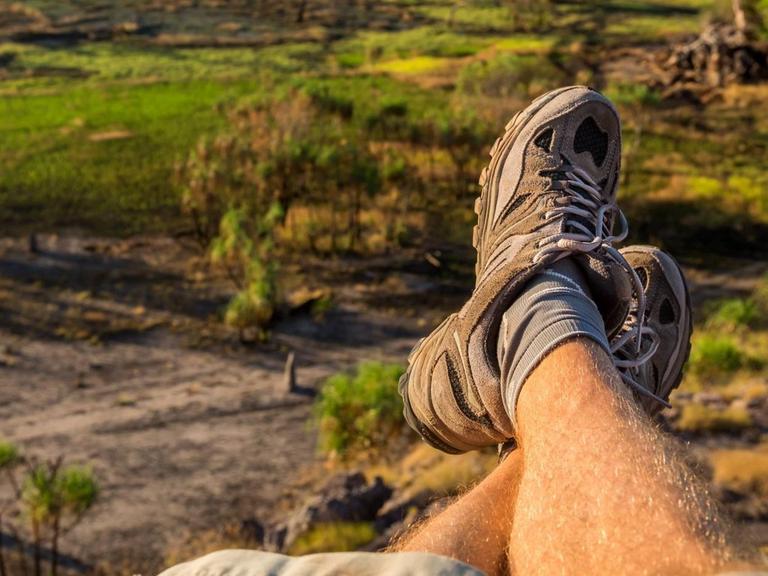 Ein Paar Männerbeine in Treckingschuhen, im Hintergrund die Nadab Floodplains in Australien.
